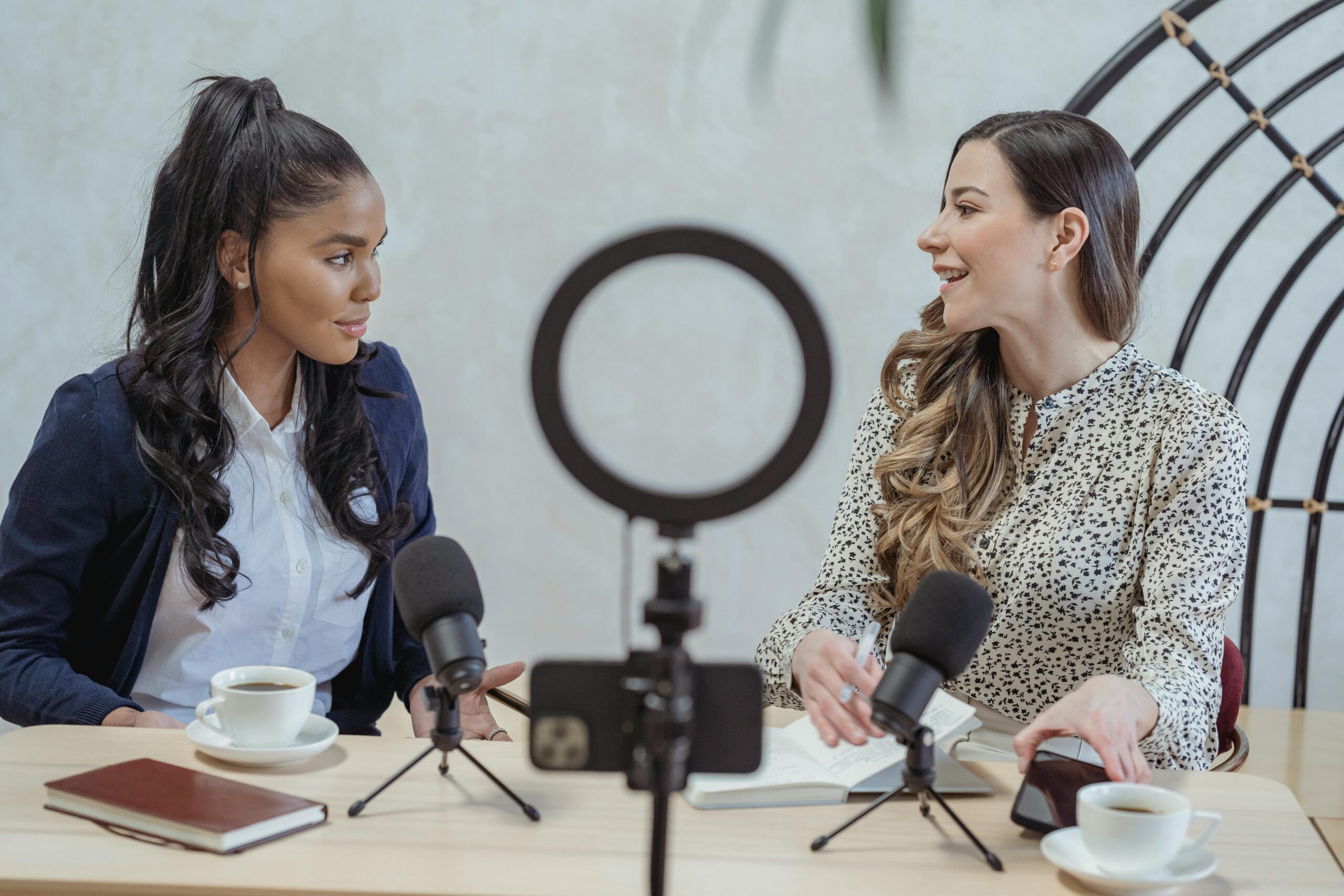 Two women talking into podcast microphones with a phone camera and ring light set up in front of them.